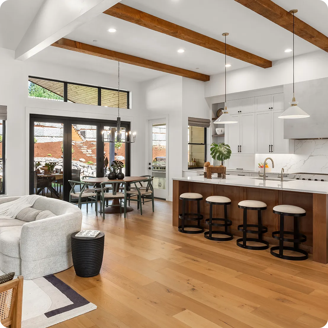 Open-concept kitchen and living area with wooden floors, white cabinets, a large island with barstools, and exposed ceiling beams. A dining table is visible near large windows.